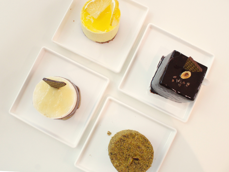 Quatro sobremesas gourmet em pratos quadrados brancos, incluindo bolo de limão amarelo, um de chocolate escuro e um com cobertura de pistache. A foto é tirada de cima, destacando a apresentação sofisticada dos doces.