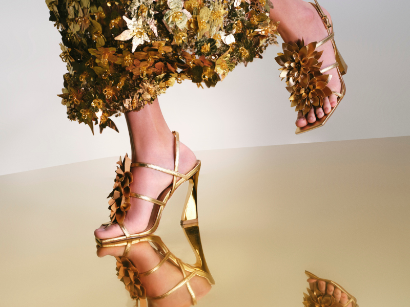 Um par de sandálias douradas com salto escultural fino e grandes flores douradas em 3D sobre o peito do pé está refletido em um chão brilhante. Um vestido luxuoso, feito de flores e folhas douradas, emoldura a parte superior da imagem.