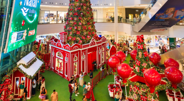 A foto mostra o cenário de Natal do RioMar Recife, com decorações predominantemente na cor vermelha, uma árvore de natal com luzes douradas, e um carrossel com balões vermelhos.