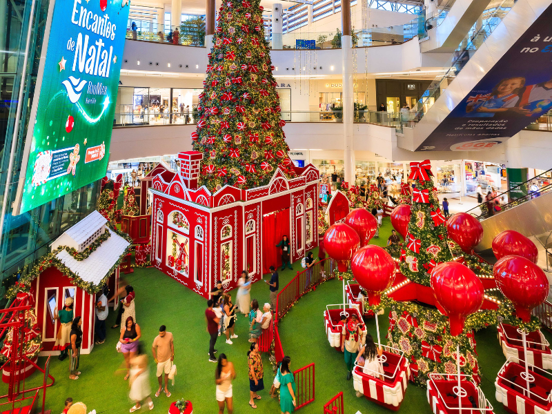 A foto mostra o cenário de Natal do RioMar Recife, com decorações predominantemente na cor vermelha, uma árvore de natal com luzes douradas, e um carrossel com balões vermelhos.