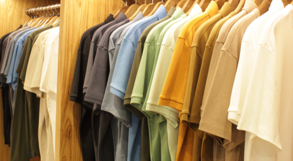 Uma se&ccedil;&atilde;o da loja com prateleiras de madeira clara, onde muitas camisetas de manga curta est&atilde;o penduradas lado a lado. As cores variam em degrad&ecirc;, do branco e cinza ao verde, mostarda e azul.