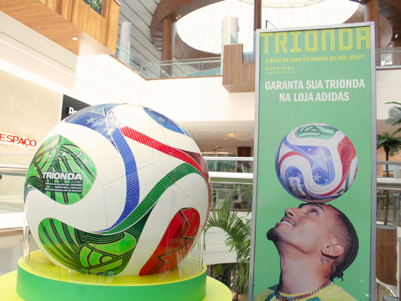 Um close-up da bola gigante Trionda em exposição, destacando seu design com as cores e padrões da Copa. A bola está sobre uma base verde-limão em um pedestal transparente.