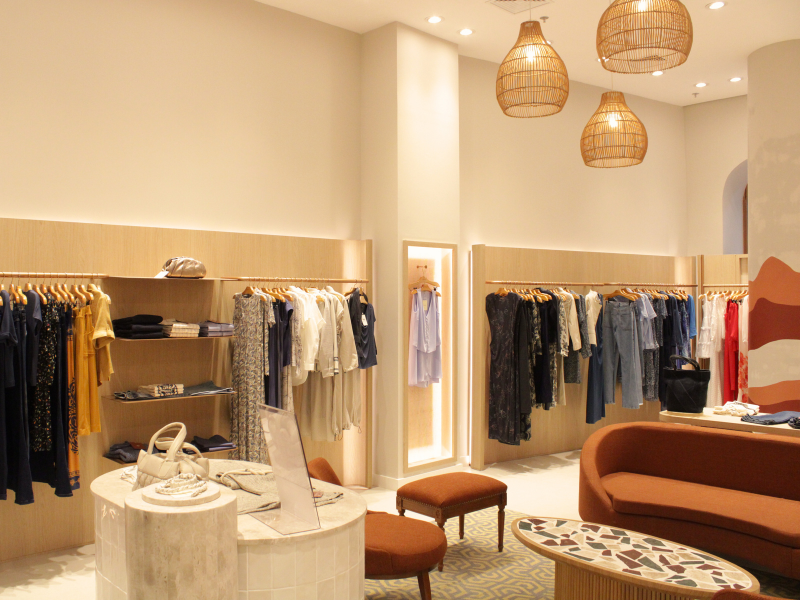 Uma seção da loja com roupas penduradas em cabides de madeira, apresentando luminárias de vime penduradas e um sofá marrom curvo.