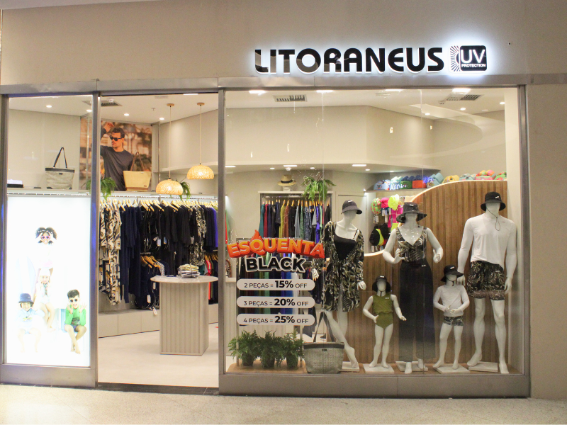 A vitrine da loja Litoraneus UV Protection exibe manequins adultos e infantis vestindo roupas de praia e proteção UV com estampa de folhagem. Um cartaz vermelho e amarelo anuncia a promoção 