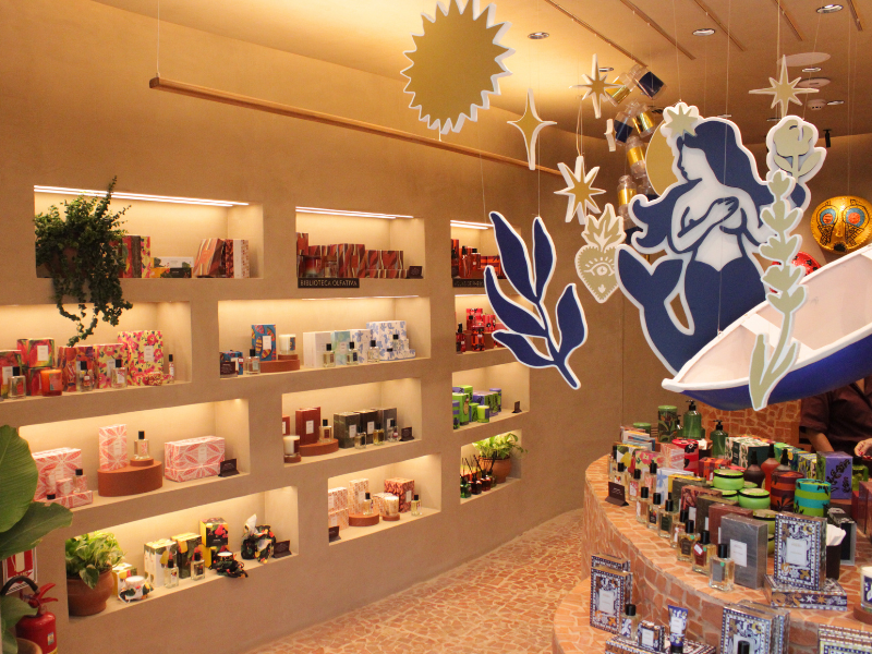 Vista interna da loja, mostrando prateleiras nichadas com produtos embalados e a decoração suspensa da sereia e barco. O piso é de azulejos rústicos em tons terrosos.