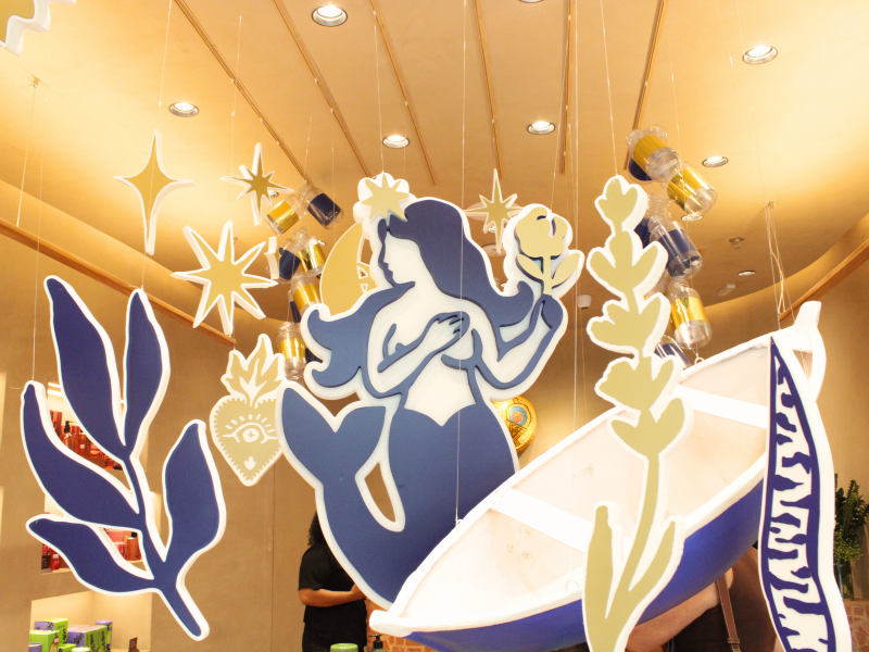 Decoração suspensa no interior da loja, mostrando recortes de papel de uma sereia, folhas, estrelas e outros elementos flutuando. A iluminação é quente.