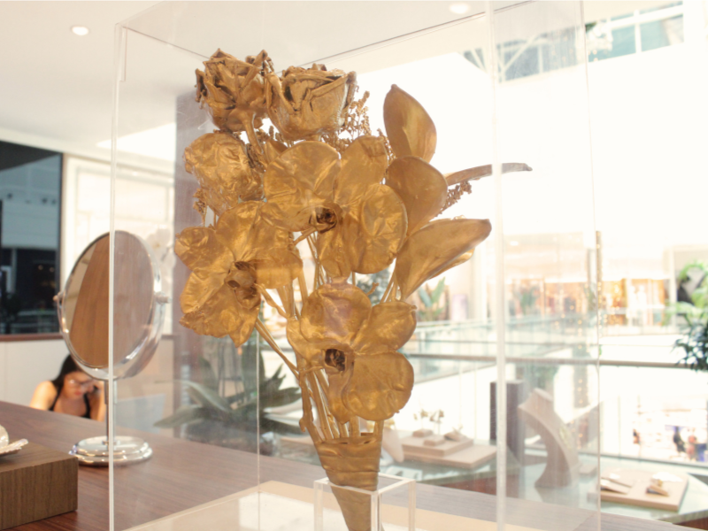 Arranjo de flores e folhas banhadas a ouro (rosas e orquídeas) em exibição dentro de uma caixa de acrílico no quiosque.