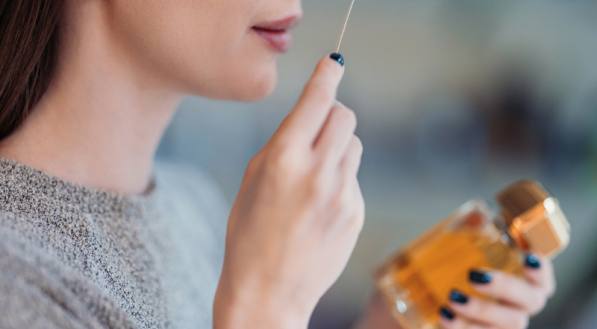 Uma mulher está provando a fragrância de um perfume importado, segurando um frasco âmbar na mão direita. Ela leva à ponta do nariz uma vareta fina de teste.
