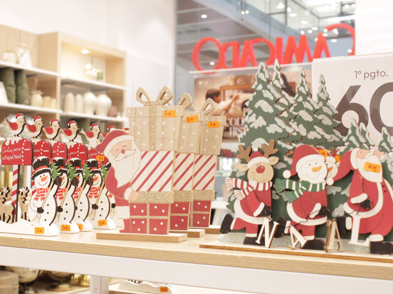 Uma exibição de decorações de Natal de madeira, incluindo bonecos de neve, Papai Noel e renas ao lado de caixas de presente brilhantes, na prateleira de uma loja.