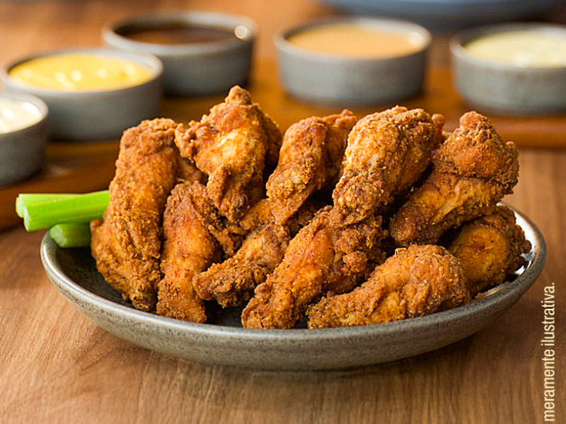 Um prato raso e cinza com uma montanha de asinhas de frango (chicken wings) empanadas e fritas, de cor dourada escura, acompanhadas por talos de aipo (salsão) à esquerda. Ao fundo, fora de foco, há vários potinhos com molhos para mergulhar.