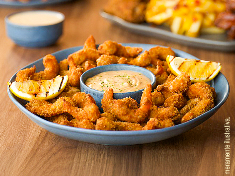Um prato azul repleto de camarões empanados e fritos, dispostos ao redor de um pequeno pote com molho cremoso ao centro. Duas fatias de limão grelhado enfeitam as laterais do prato.
