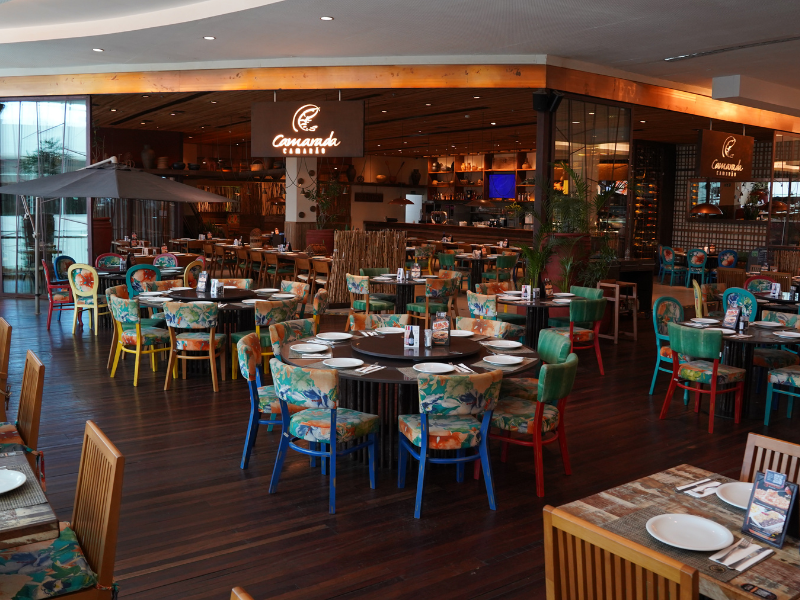 O amplo salão do restaurante Camarada Camarão, com piso de madeira escura, mesas redondas e cadeiras coloridas com estampas florais. O nome do restaurante brilha em um letreiro sobre o balcão.
