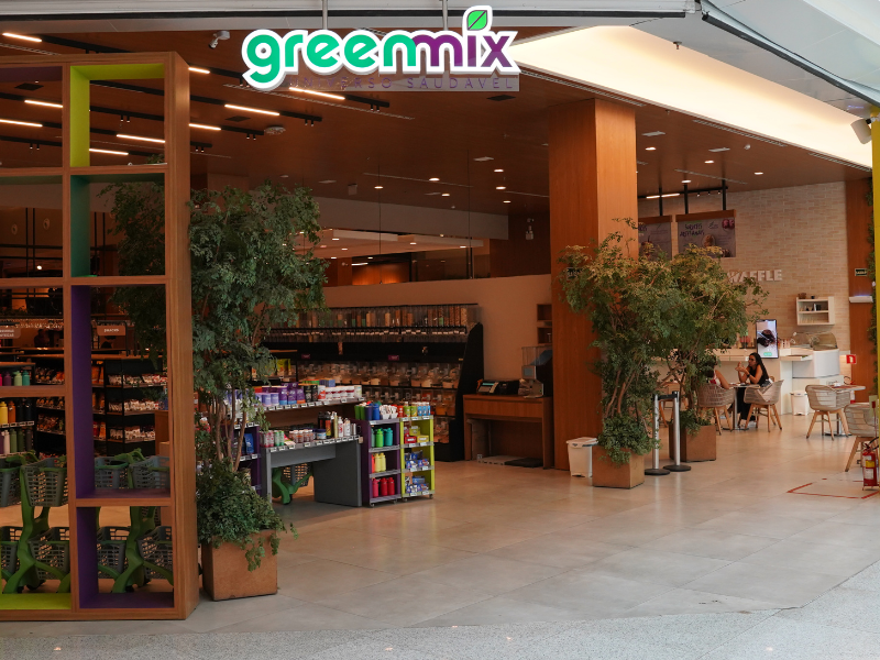 A entrada da loja Greenmix, com caixas registradoras e prateleiras de produtos saudáveis ao fundo. A loja tem um design em madeira com grandes vasos de plantas e carrinhos de compras verdes.