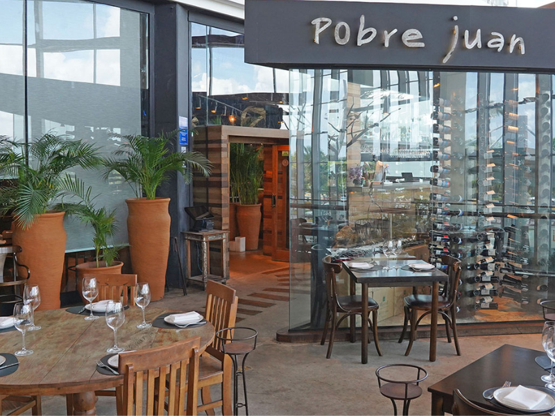 A fachada externa e envidraçada do restaurante Pobre Juan, mostrando mesas e cadeiras de madeira e uma grande prateleira de vinhos no interior. Grandes vasos de barro com palmeiras decoram a área.