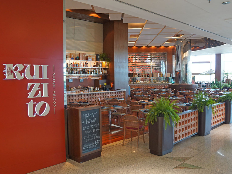 A entrada do restaurante Ruizito - Cocina Ibérica, com uma parede vermelha e o letreiro da marca. Uma lousa com o menu de "Happy Hour" está na entrada, e o salão tem decoração em tons de madeira e marrom.