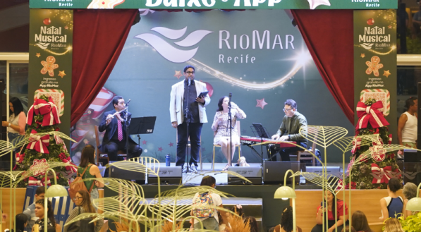 Palco do evento "Natal Musical RioMar Recife", com uma banda e cantores se apresentando sob cortinas vermelhas e um banner de Natal, com o público assistindo na frente.