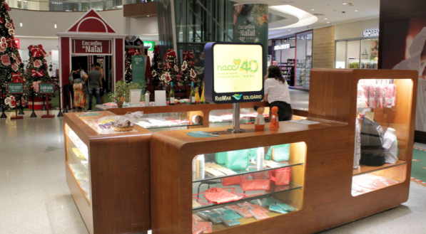Vista geral de um quiosque de madeira em um shopping, vendendo produtos artesanais, com uma tela de TV exibindo a logomarca "Nacc 40 anos" e uma decoração de Natal ao fundo.