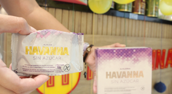 A imagem mostra uma mão segurando lado a lado um alfajor Havanna "Sin Azúcar" (Sem Açúcar), embalado individualmente (70g) e uma caixa de alfajor Havanna "Sin Azúcar" (Sem Açúcar), contendo 4 unidades.