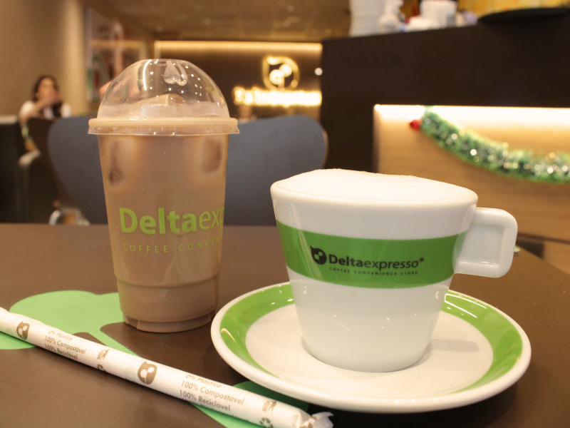 Um cappuccino quente em xícara branca com detalhes verdes e uma bebida gelada no copo com logo da Deltaexpresso, sobre uma mesa com um canudo compostável.