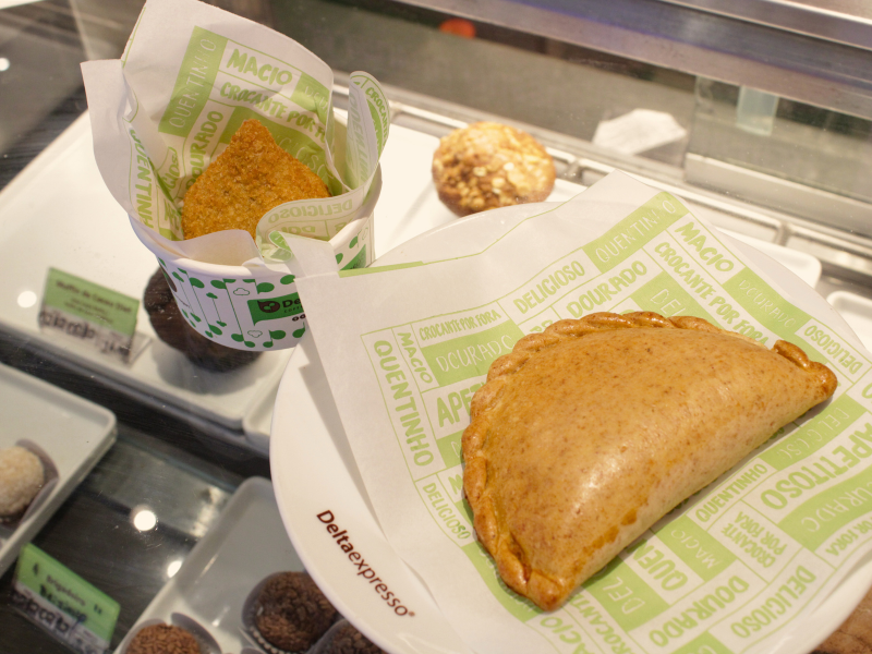 Visão de perto de salgados e doces em uma vitrine da Deltaexpresso, destacando uma coxinha em copo de papel e uma empanada em prato com guardanapo temático.