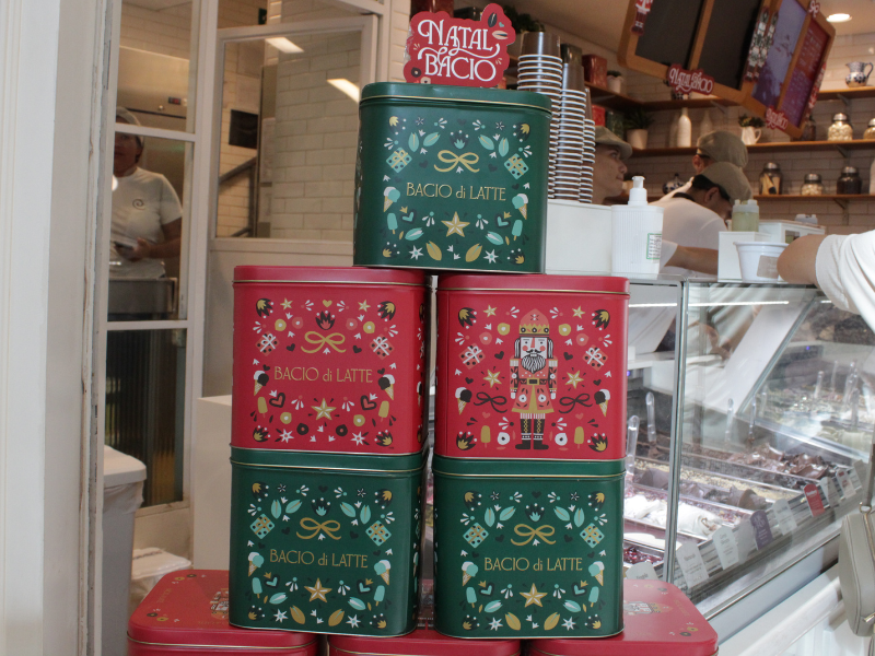 Pilha de latas decorativas de Natal da marca Bacio di Latte, em cores vermelho e verde, com temas de quebra-nozes e sorvetes.