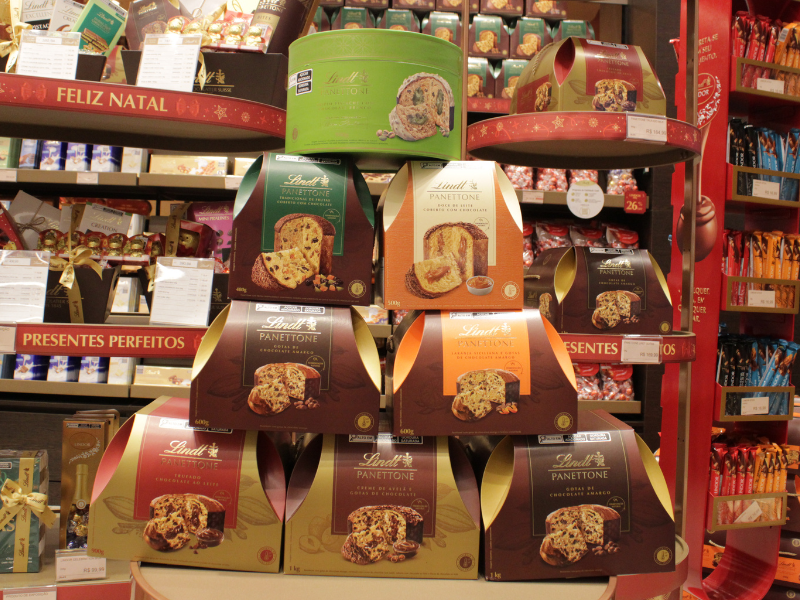 Exposição de vários Panetones Lindt em caixas de cores variadas, organizados em uma prateleira de loja, com alguns preços visíveis ao fundo.