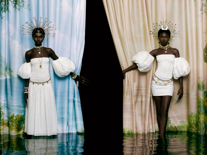 Duas modelos posam em um cenário de cortinas azuis e bege sobre água, vestindo roupas brancas e volumosas. Ambas usam joias extravagantes, coroas douradas e unhas longas.