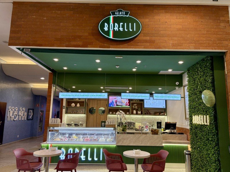 Fachada da loja Getalo Borelli com testeira com tijolos cor marrom, loja internamente pintada de verde, com duas mesas pequenas e duas cadeiras em cada. Um vitrine com opções de gelatos e caixa para registro de compras.