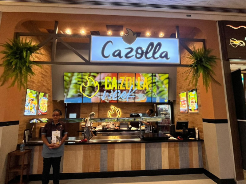 Fachada da nova loja Cazolla localizada no L3, praça de alimentação com duas atendentes à frente posando para foto.