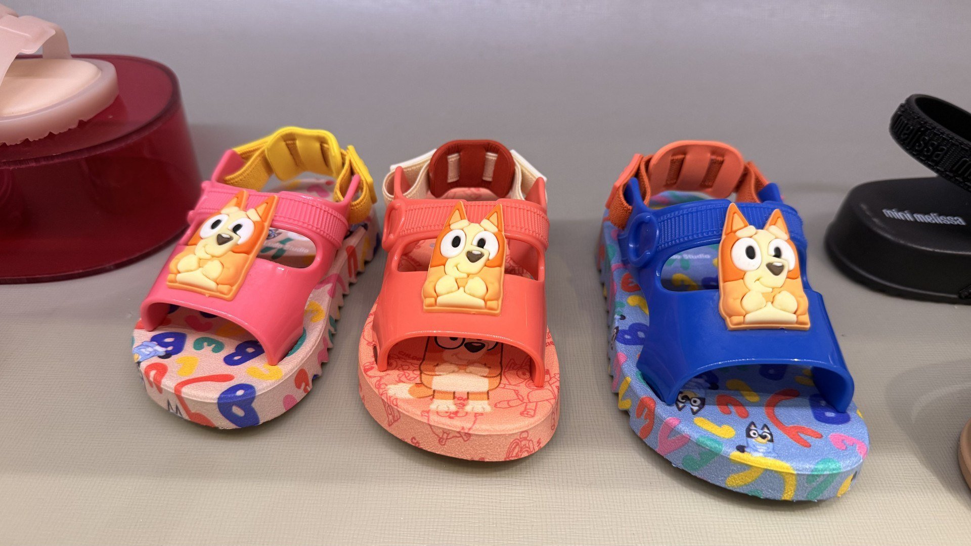 Três sandálias da Melissa com o tema de Bluey, no valor de 179,90 reais cada. Uma sandália rosa, outra laranja e outra azul. 