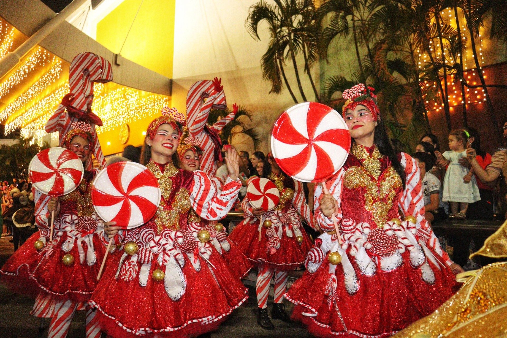 Mulheres vestidas de bengalinha de Natal e com pirulitos grandes. Elas sorriem para as pessoas que acompanham o desfile.  Mulheres vestidas de bengalinha de Natal e com pirulitos grandes. Elas sorriem para as pessoas que acompanham o desfile.