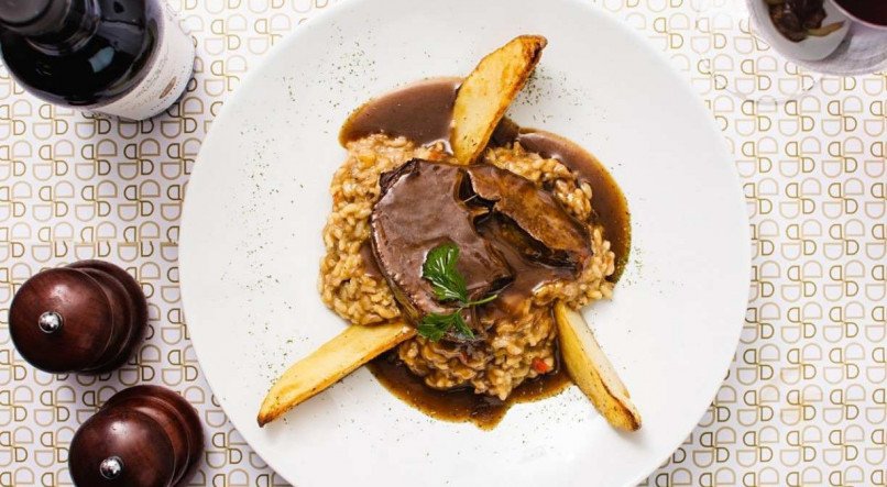Cordeiro Douro In: pernil de cordeiro marinado em vinho, especiarias e ervas frescas, assado lentamente em forno combinado, servido em seu próprio molho sobre risoto das lascas de cordeiro e batatas rústicas