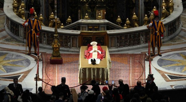 Bento XVI deitado sobre um catafalco, vestido de branco com uma casula vermelha, cor do luto papal, com uma mitra branca debruada a ouro e um ros&aacute;rio entrela&ccedil;ado nas m&atilde;os