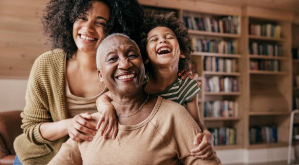 Imagem de m&atilde;e, filha e neta felizes, em frente a uma estante repleta de livros.