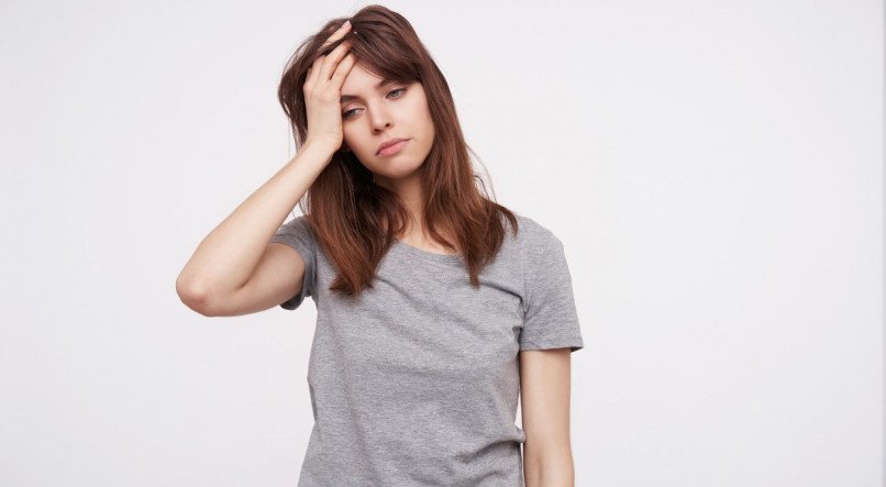 Indoor shot of young pretty brunette female holding raised hand on her head and looking tiredly down while standing over white background in casual clothes