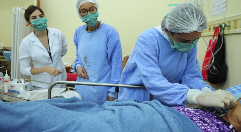 Enfermeiras com toucas, m&aacute;scaras e aventais enquanto realizam procedimento em paciente. 
