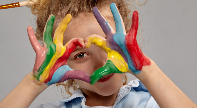 Close-up shot of a funny little girl having a brush in her chic curly blond hair, wearing in a blue shirt and white t-shirt. She is looking through a painted hands folded in the shape of a heart and smiling, on a gray background / Autismo