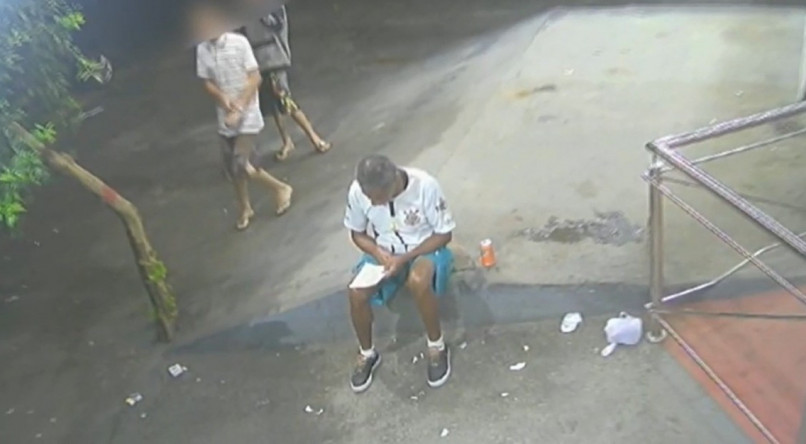 Imagens de uma câmera de segurança mostram o momento em que o idoso, sentado e concentrado nas palavras-cruzadas, é atingido pelo disparo