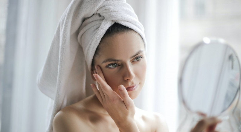 Mulher com os cabelos enrolados na toalha segurando o rosto com a pele limpa enquanto se olha no espelho.
