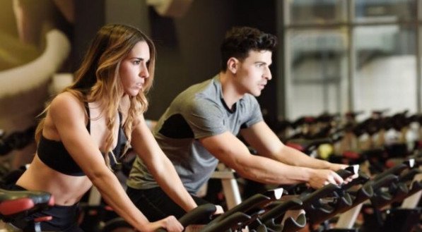 Imagem ilustrativa: homem e mulher realizam exerc&iacute;cio cardio na bike, durante treino em academia. 