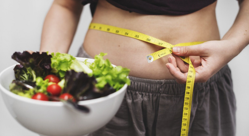 Pessoa em dieta ou processo de emagrecimento usando fita m&eacute;trica na barriga enquanto segura uma tigela de salada.