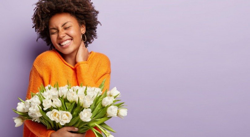 Jubilant upbeat black woman has curly hair, broad shining smile, keeps hand on neck, eyes shut, wears orange sweater, holds white flowers with pleasant scent, models over purple studio wall. Spring