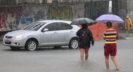 Imagem: duas mulheres de costas atravessam avenida alagada segurando guarda-chuvas; há um carro prata ao fundo