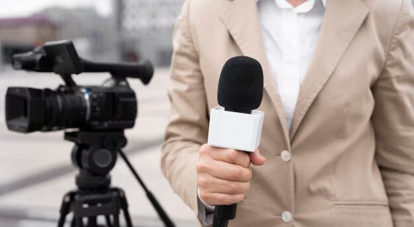 Imagem ilustrativa de uma jornalista segurando um microfone próximo a uma câmera de filmagem