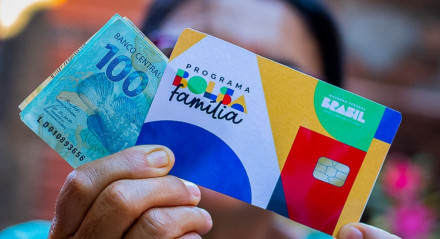Imagem ilustrativa de uma mulher segurando o cartão do Bolsa Família e notas de dinheiro