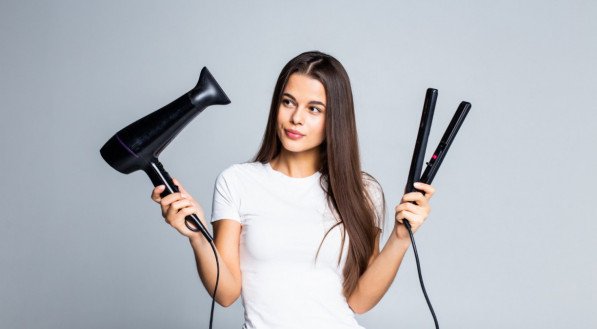 Imagem de uma mulher segurando um secador e uma chapinha, fontes de calor que danificam o cabelo com o tempo