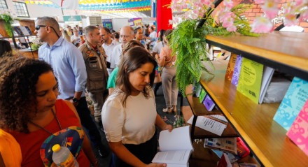 Raquel Lyra no Circuito Literário de Pernambuco em Caruaru. Governadora segura um livro aberto em frente a uma estante com diversos livros em um espaço com a bandeira de Pernambuco pendurada no teto