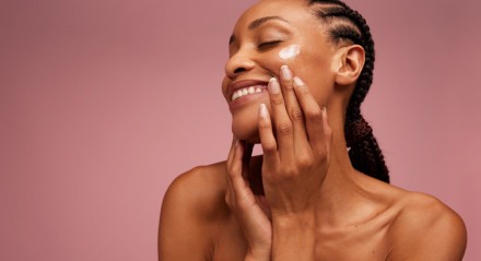 Imagem de mulher jovem com olhos fechados e sorrindo, passando hidratante no rosto; skin care