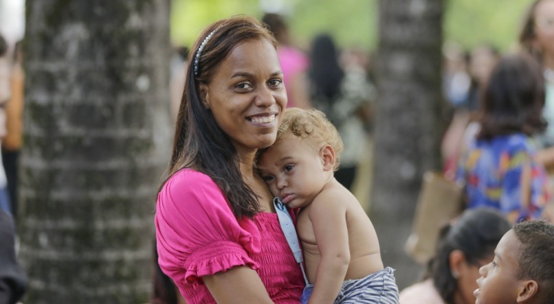 Mulher com uma criança no colo em um parque, mãe sorri para câmera enquanto a criança repousa em seu ombro