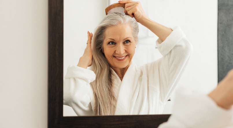 Imagem de senhora com cabelos grisalhos longos sorrindo ao se pentear na frente do espelho